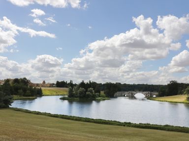 A large lake with an island and a stone bridge with arched openings in a parkland setting with trees and buildings in the distance at Blenheim Edge The Causeway Woodstock
