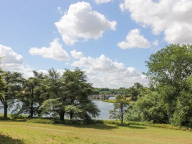 A grassy area with trees near a river and a stone bridge under a partly cloudy sky at Blenheim Edge The Causeway in Woodstock