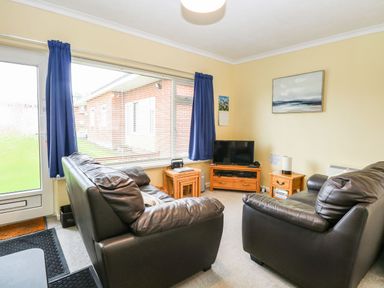 A living room with two leather sofas a TV on a wooden stand and a large window with blue curtains at 21 Siesta Mar in Mundesley