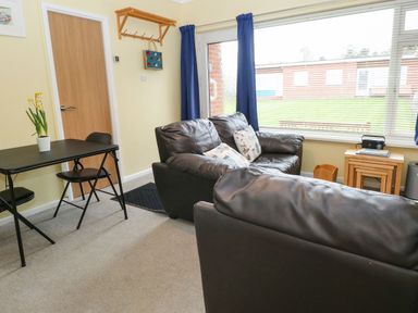A living room with two black sofas a small dining table with two chairs and a window with blue curtains at 21 Siesta Mar in Mundesley