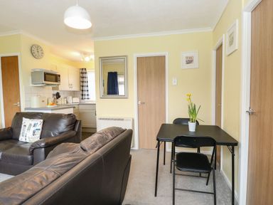 A living area with two brown sofas a black dining table with chairs and a kitchen area at 21 Siesta Mar in Mundesley