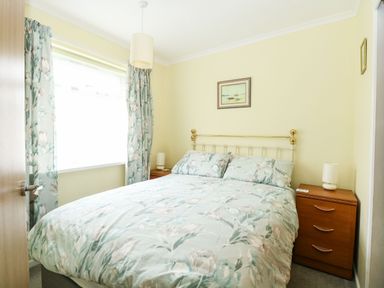 A bedroom with a double bed floral bedding two wooden bedside tables with lamps and a window with floral curtains at 21 Siesta Mar in Mundesley