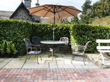 An outdoor patio area with a round table four metal chairs and a large umbrella surrounded by greenery at The Coach House in Llanrwst