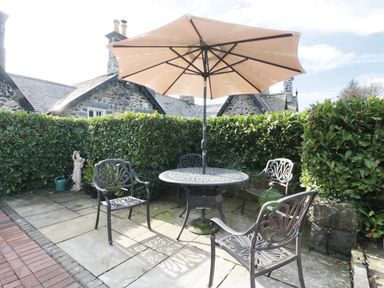 A patio area with a round metal table and four chairs under a large umbrella surrounded by hedges at The Coach House in Llanrwst