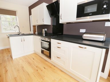 A kitchen with white cabinets black countertop built in oven microwave and stainless steel toaster at The Coach House in Llanrwst