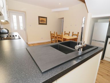 A kitchen area with a black countertop sink, wooden dining table with chairs, refrigerator and white door at The Coach House in Llanrwst