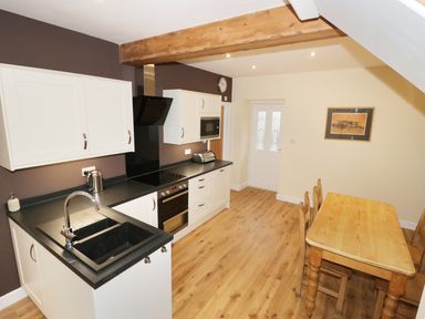 A kitchen with white cabinets black countertops a wooden dining table and chairs wood flooring and a wooden beam at The Coach House in Llanrwst