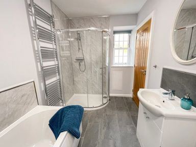 A bathroom with a bathtub and blue towel a glass shower enclosure a sink with soap dispenser and a mirror a wooden door and a window at The Coach House in Llanrwst