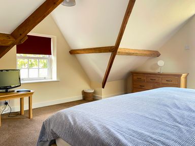 A bedroom with a bed covered in a striped duvet a wooden dresser a small wooden table with a television and a window with a red blind at The Coach House in Llanrwst