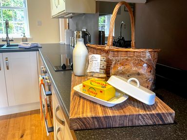 A kitchen countertop with a cutting board holding a bottle of milk butter in a dish a pack of welsh cakes and a loaf of bread next to a wicker basket at The Coach House in Llanrwst