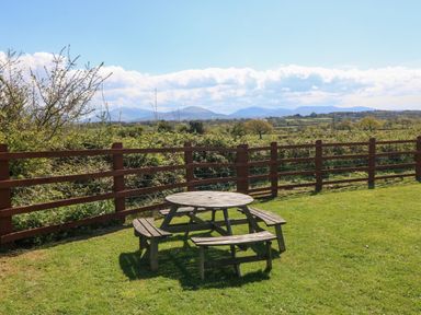 A round wooden picnic table and benches on grass near a wooden fence with trees and mountains in the distance at Ty Mawr in Llanddaniel Fab