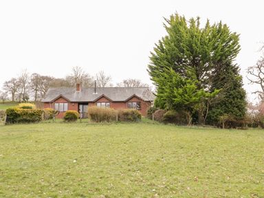 A single-story brick house with a sloped roof behind bushes and trees on a grassy field at The Alders in Llanfyllin