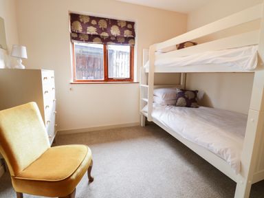 A bedroom with white bunk beds a yellow chair a white chest of drawers and a window with patterned blinds at The Alders in Llanfyllin