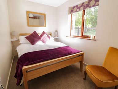 A bedroom with a wooden bed white bedding and purple pillows beside a window and a mustard chair at The Alders in Llanfyllin