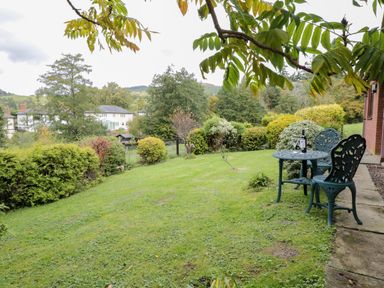 A garden with green grass and bushes and a metal table and two chairs with wine bottle and glasses at The Alders in Llanfyllin