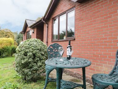 A metal outdoor table with two chairs and a bottle of wine and two glasses next to a brick house at The Alders in Llanfyllin