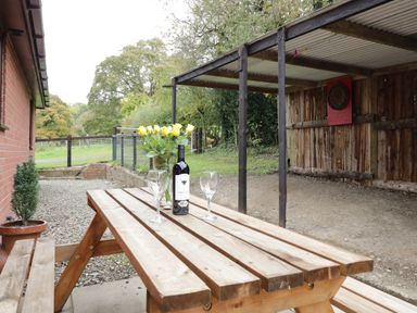 A wooden picnic table with two wine glasses a bottle of wine and yellow flowers near a brick wall and a covered wooden area at The Alders in Llanfyllin