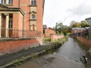 A narrow water canal flowing beside brick buildings and a paved path at The Alders in Llanfyllin