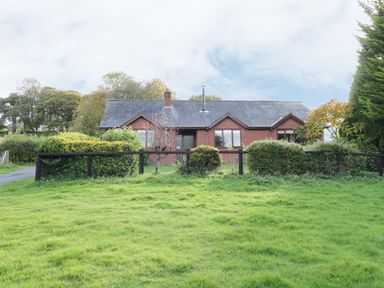 A single-story brick house with a slate roof behind a black fence and bushes at The Alders in Llanfyllin