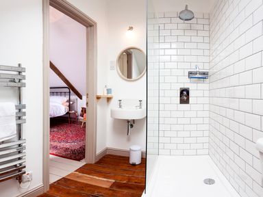 A bathroom with a shower and sink with a round mirror and wooden floor leading to a bedroom with a bed and red carpet at Star House in Marazion