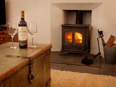 A room with a wood burning stove firewood bucket fireplace tools and a table with a wine bottle and two glasses at Star House in Marazion