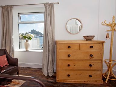 A room with a wooden chest of drawers a round mirror a vase with flowers on the windowsill a coat rack and a chair at Star House in Marazion