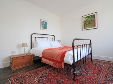 A bedroom with a metal bed frame red patterned rug wooden chest as a bedside table and two framed pictures on white walls at Star House in Marazion