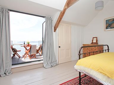 A bedroom with a metal bed frame and yellow blanket next to a wooden dresser and open doors leading to a balcony with a wooden table and chairs at Star House in Marazion