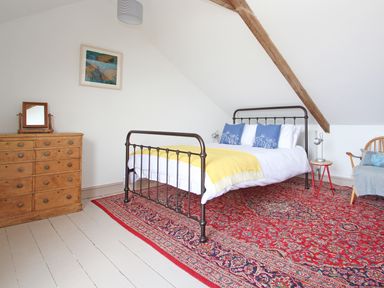 A bedroom with a metal bed frame white bedding and yellow blanket on a red patterned rug with a wooden dresser and chair at Star House in Marazion