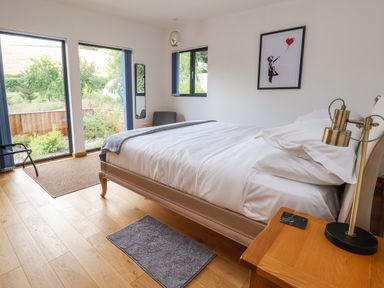 A bedroom with a bed white bedding wooden floor side table gold lamp and windows showing greenery at No1 The Lodge in Acton near Bishops Castle