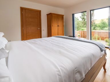 A bedroom with a bed white bedding wooden wardrobe wooden door and large windows showing a garden at No1 The Lodge in Acton near Bishops Castle