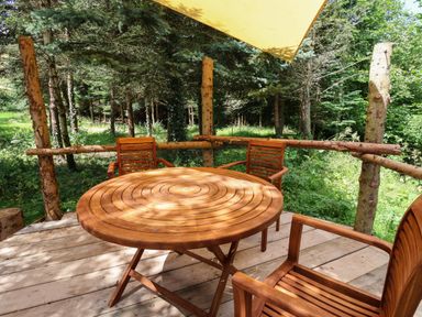 An outdoor wooden deck with a round wooden table and three wooden chairs surrounded by trees at No1 The Lodge in Acton near Bishop's Castle