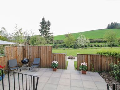 A patio with two chairs a barbecue and potted plants overlooking a grassy lawn with trees and hills at No1 The Lodge in Acton near Bishops Castle