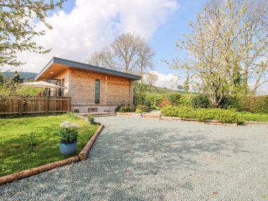 A wooden building with gravel driveway and garden with trees and plants at No1 The Lodge in Acton near Bishops Castle