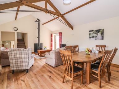 A dining area with a wooden table and six chairs next to a living room with sofas a plaid armchair a wood stove and exposed wooden beams at Pentre Fach in Llandrindod Wells