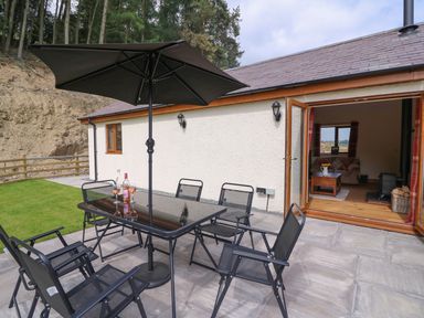 An outdoor patio with a glass table umbrella and chairs next to a house with open doors showing a living room at Pentre Fach in Llandrindod Wells