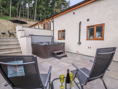 An outdoor patio area with two chairs and a small glass table with a bottle and two glasses a hot tub and stairs leading to a higher level at Pentre Fach in Llandrindod Wells