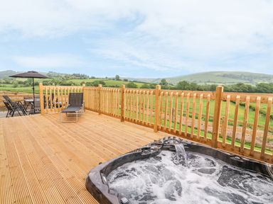 An outdoor wooden deck with a hot tub a lounge chair and a table with chairs and umbrella overlooking green hills at Pentre Fawr in Llandrindod Wells