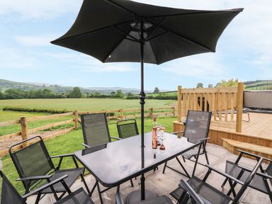 An outdoor patio table with six chairs and a large umbrella overlooking a fenced grassy field and hills at Pentre Fawr in Llandrindod Wells