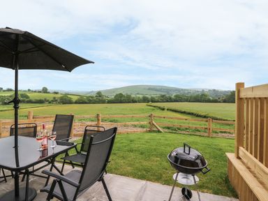 A patio area with a table and four chairs a bottle of wine and two glasses a barbecue and a wooden fence overlooking fields and hills at Pentre Fawr in Llandrindod Wells