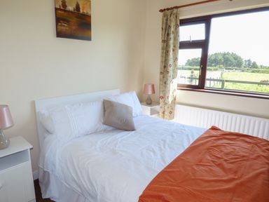 A bedroom with a bed white pillows an orange blanket two bedside tables with lamps a window with floral curtains and a painting on the wall at Nephin View in Foxford County Mayo