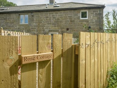 A wooden fence gate with a sign reading Rose Cottage and a stone building in the background at Rose Cottage in Edlingham