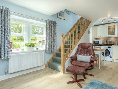 A living area with a brown leather chair and footrest near stairs and a small kitchen with appliances at Rose Cottage in Edlingham