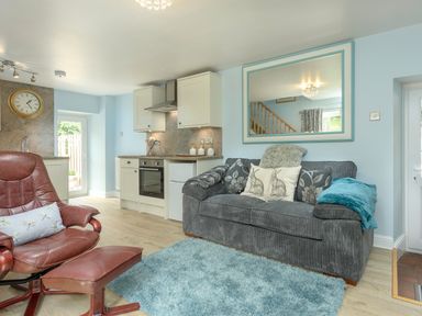 An open plan living area with a gray sofa a blue rug a brown leather chair and footstool and a kitchen with white cabinets at Rose Cottage in Edlingham