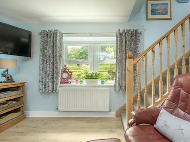 A living room window with grey curtains a wooden staircase a brown leather chair a wooden cabinet with a lamp and decorative items at Rose Cottage in Edlingham