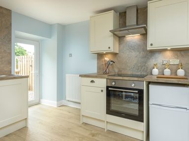 A kitchen with built in oven hob cabinets a small fridge and door to outside at Rose Cottage in Edlingham