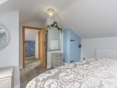 A bedroom with a bed covered in patterned bedding a small dresser a mirror a wooden door and a blue slanted closet door at Rose Cottage in Edlingham