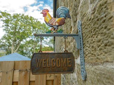 A metal welcome sign with a colorful rooster mounted on a stone wall outside Rose Cottage in Edlingham