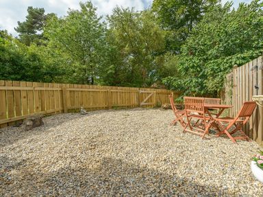 A gravel patio with wooden table and chairs and a wooden fence surrounded by trees at Rose Cottage in Edlingham