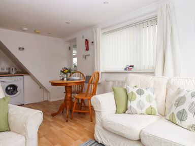 A living area with a white sofa with leaf-patterned cushions wooden dining table and chairs and a washing machine under a staircase at Wisteria Cottage in Exmouth